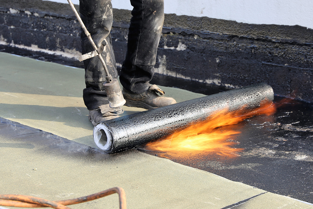 A construction professional applying a torch-on waterproofing membrane to a flat roof. A propane torch is being used to melt the asphalt roll as it is applied to the surface.