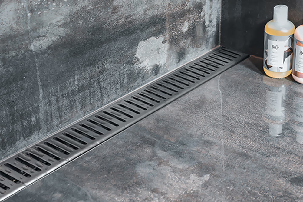 A close-up of a modern walk-in shower featuring a sleek, stainless steel linear drain installed along the base of a dark, textured tile wall. The floor consists of large-format grey stone tiles with a wet, reflective surface, and two bottles of hair care products are visible in the corner.