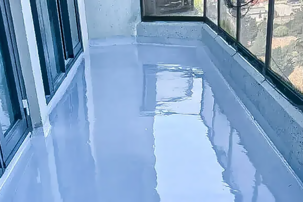 A long balcony featuring a freshly applied, high-gloss light blue waterproof coating on the floor. The reflective surface mirrors the surrounding glass windows and balcony railings, showing a professional waterproofing or floor finishing project in progress.